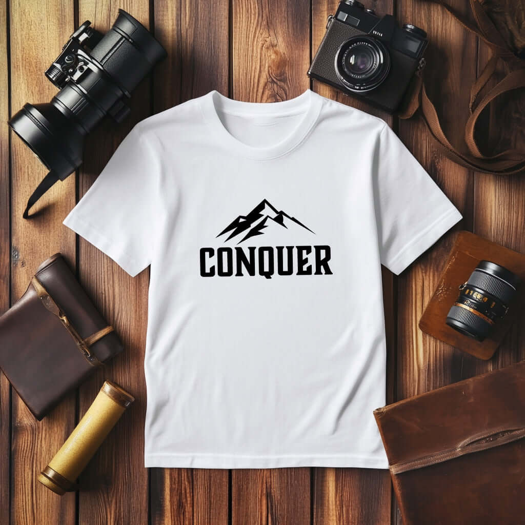 Conquer Men's White T-shirt displayed on a wooden surface with cameras and accessories.