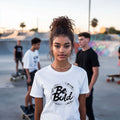 Young woman wearing Be Bold unisex white t-shirt, standing in skate park with friends, inspired confidence and individuality.